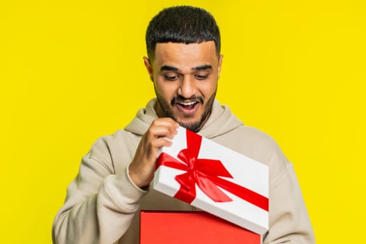 Happy Indian young man opening birthday gift box with red ribbon. Holidays surprise concept. Smiling Arabian Hindu guy receive great wrapped present celebrating isolated on yellow studio background