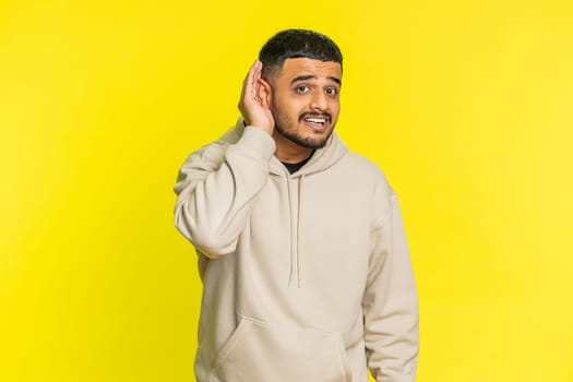 I cant hear you. What. Indian man trying hear you, looking confused and frowning, keeping arm near ear for louder voice, asking to repeat, to hear information, deafness. Hindu guy on yellow background