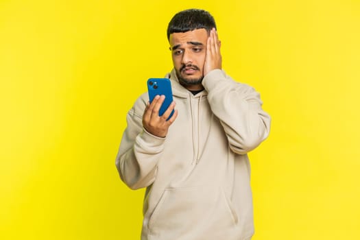 Upset sad Indian man use smartphone typing browsing, play game, loses, surprised by sudden lottery results bad fortune loss fail unlucky news. Arabian Hindu bearded guy isolated on yellow background