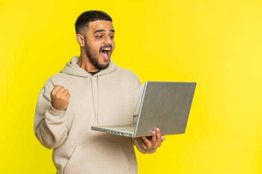 Excited happy Indian man typing on laptop computer, working on project, enjoying results, winning lottery game, celebrating success, online shopping, victory. Arabian guy isolated on yellow background