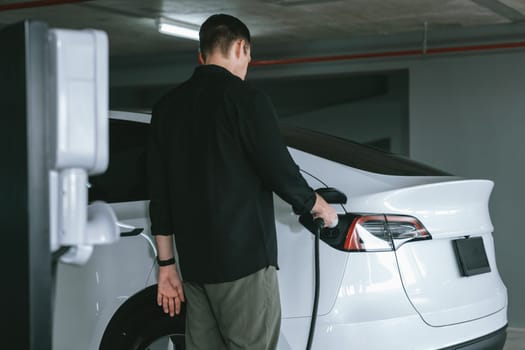 Young man travel with EV electric car to shopping center parking lot charging in downtown city showing urban sustainability lifestyle by green clean rechargeable energy of electric vehicle innards