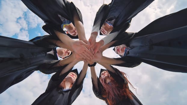 Team of college or university students celebrating graduation. Group of happy successful graduates in academic hats and robes standing in circle and putting their hands together