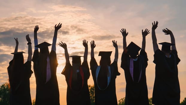 Silhouettes of graduates in black robes waving their arms against the evening sunset