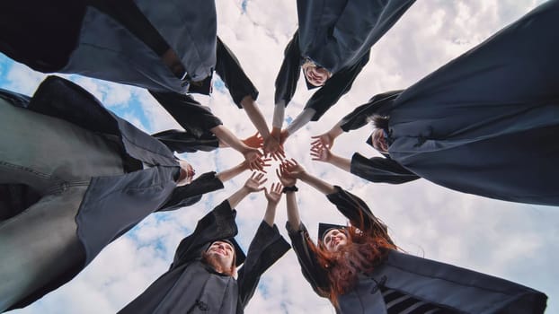 Team of college or university students celebrating graduation. Group of happy successful graduates in academic hats and robes standing in circle and putting their hands together