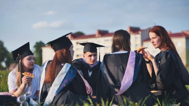 Graduates in black suits eating pizza in a city meadow