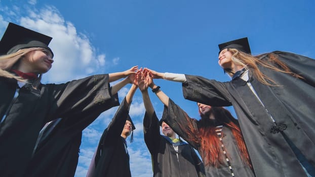 Team of college or university students celebrating graduation. Group of happy successful graduates in academic hats and robes standing in circle and putting their hands together.