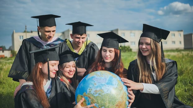 Graduates in black robes examine a geographical globe sitting on the grass