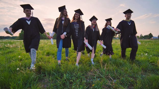 Six graduates in robes walk against the backdrop of the sunset