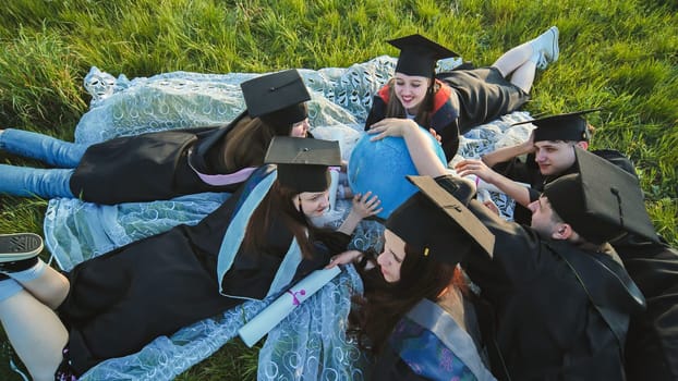 Graduate students in black robes study a globe on the grass