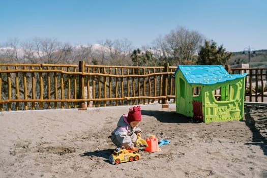 Little girl digs sand on the playground in the park near the toy house. High quality photo