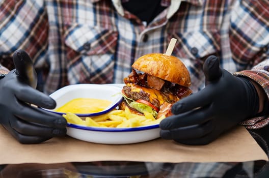 The concept of fast food and takeaway food. A man in black latex gloves is ready to eat a juicy hamburger that lies near the french fries on a metal plate along with cheese sauce