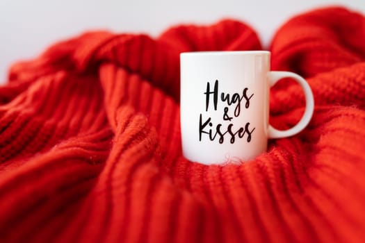 A white cup with a heart stands on a red knitted fabric. Inscription of St. Valentine's day, 14 February