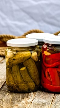 Glass jars with pickled red bell peppers and pickled cucumbers (pickles) isolated. Jars with variety of pickled vegetables. Preserved food concept in a rustic composition.