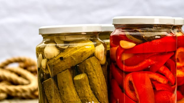 Glass jars with pickled red bell peppers and pickled cucumbers (pickles) isolated. Jars with variety of pickled vegetables. Preserved food concept in a rustic composition.