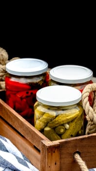 Wooden crate with glass jars with pickled red bell peppers and pickled cucumbers (pickles) isolated. Jars with variety of pickled vegetables. Preserved food concept in a rustic composition.