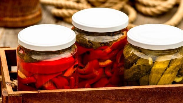 Wooden crate with glass jars with pickled red bell peppers and pickled cucumbers (pickles) isolated. Jars with variety of pickled vegetables. Preserved food concept in a rustic composition.