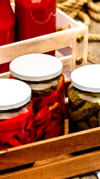 Wooden crate with glass jars with pickled red bell peppers and pickled cucumbers (pickles) isolated. Jars with variety of pickled vegetables. Preserved food concept in a rustic composition.