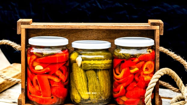 Wooden crate with glass jars with pickled red bell peppers and pickled cucumbers (pickles) isolated. Jars with variety of pickled vegetables. Preserved food concept in a rustic composition.