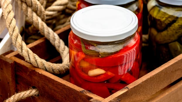 Wooden crate with glass jars with pickled red bell peppers and pickled cucumbers (pickles) isolated. Jars with variety of pickled vegetables. Preserved food concept in a rustic composition.