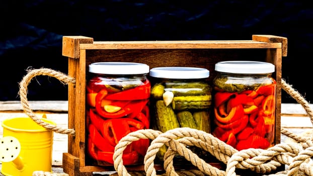 Wooden crate with glass jars with pickled red bell peppers and pickled cucumbers (pickles) isolated. Jars with variety of pickled vegetables. Preserved food concept in a rustic composition.