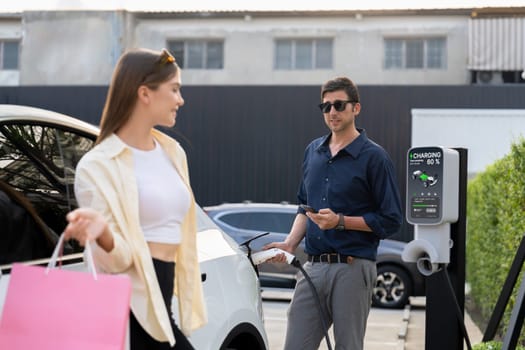 Young couple holding shopping bag and use smartphone to pay for electricity for recharging EV car battery from charging station at city mall parking lot. Happy couple go shopping by eco car. Expedient