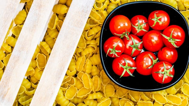Top view of pasta in a wooden crate and fresh ripe cherry tomatoes in a black bowl. Ingredients and food concept