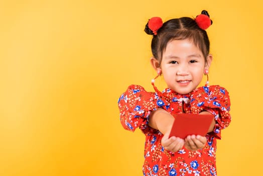Chinese New Year. Happy Asian Chinese little girl smile wear red cheongsam handed angpao or red packet monetary gift, Portrait children in traditional dress, studio short isolated on yellow background