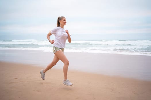 Woman running along ocean shore beach. Jogging on nature Woman in white shirt top and green sports shorts runs along seashore. Athletic woman run along sandy beach on seashore in morning
