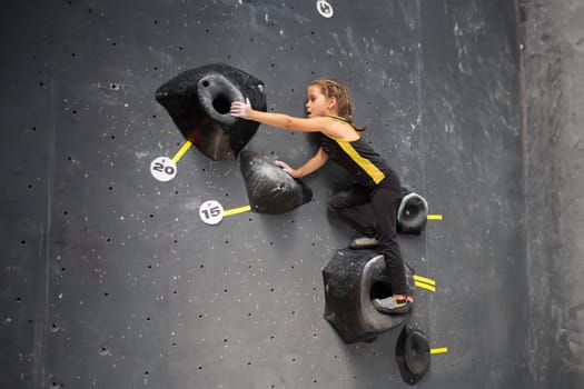 Little girl trained climbs wall at climbing centre. Child with brown hair in sports clothes working out in children's bouldering centre without assistance. Brave and strong girl climbs up mock wall