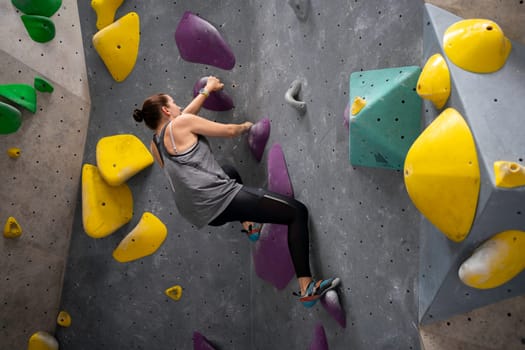 Mature woman climbs an exercise mountain at gym. Adult woman practicing in climbing wall. Woman practicing bouldering in sports center, climbing colored artificial wall without safety rope
