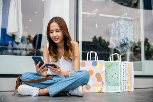 Millennial woman enjoying retail therapy, sitting on the floor with shopping bags, credit card, and smartphone in hand. Urban store window in the background. Shopping is her relaxation.