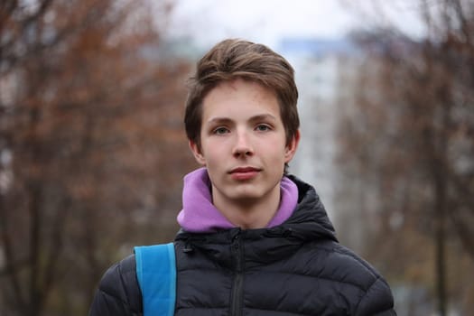 Close-up portrait of a smart and positive boy 14-17 years old in a black jacket against the background of autumn trees without leaves, bokeh.