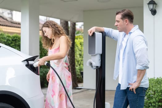 Happy and lovely couple with eco-friendly conscious recharging electric vehicle from EV home charging station. EV car technology utilized for residential home to future sustainability. Synchronos