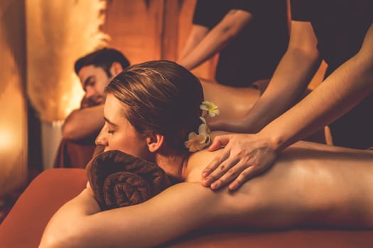 Caucasian couple customer enjoying relaxing anti-stress spa massage and pampering with beauty skin recreation leisure in warm candle lighting ambient salon spa at luxury resort or hotel. Quiescent