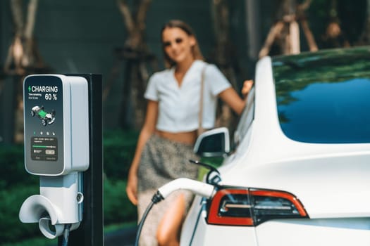 Young woman travel with EV electric car charging in green sustainable city outdoor garden in summer. Urban sustainability lifestyle by green clean rechargeable energy of electric BEV vehicle innards