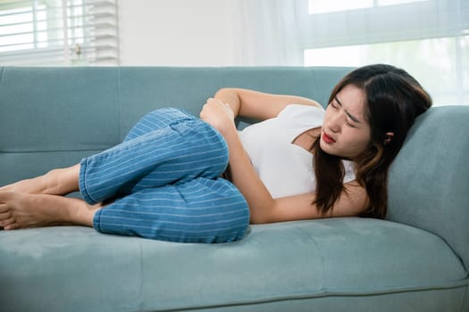 Sad Asian woman close eyes she problem painful stomachache, young female unhappy hands holding on stomach suffering from abdominal pain on sofa at home in living room, medical care and health concept