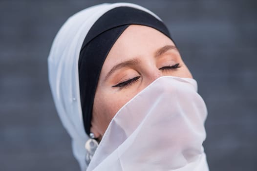 Portrait of a young woman in a hijab against a gray brick wall. Muslim woman with white scarf covering her face