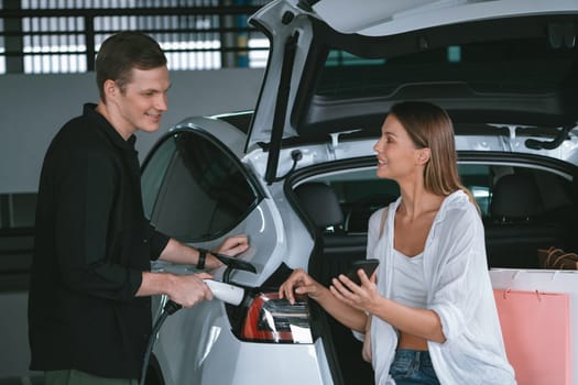 Young couple travel with EV electric car to shopping center parking lot charging in downtown city showing urban sustainability lifestyle by green clean rechargeable energy of electric vehicle innards