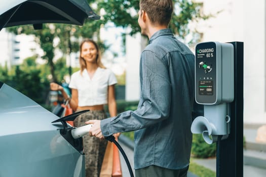 Young couple travel with EV electric car charging in green sustainable city outdoor garden in summer shows urban sustainability lifestyle by green clean rechargeable energy of electric vehicle innards