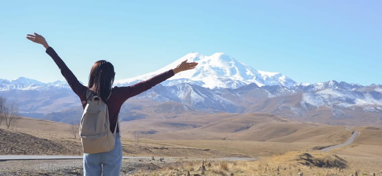 A happy female traveler with a backpack behind her back and her arms raised up admires the snow-covered Elbrus on a clear autumn day. View of Elbrus, North Caucasus, Russia.