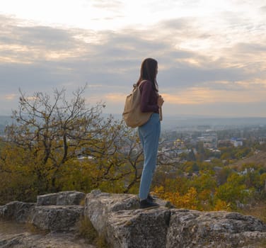 A girl with a backpack on top of a mountain looks at the city below at sunset in autumn. Hiking. Nature.