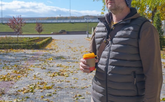 A young man walks in an autumn park and drinks coffee, rests, relaxes. Urban lifestyle concept