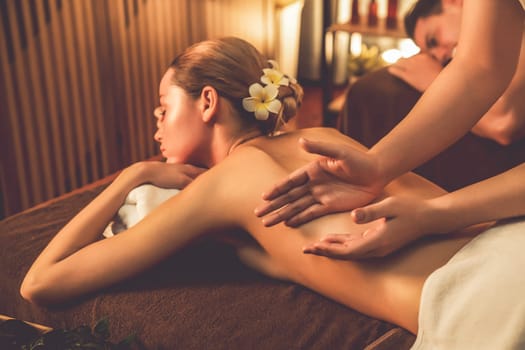 Caucasian couple customer enjoying relaxing anti-stress spa massage and pampering with beauty skin recreation leisure in warm candle lighting ambient salon spa at luxury resort or hotel. Quiescent