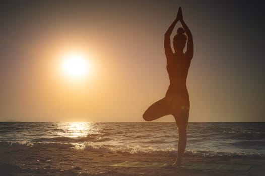 Full length rear view of a silhouette of a woman standing on one leg while practicing tree pose in yoga on a tranquil beach, taken at sunset or sunrise during summer vacation.