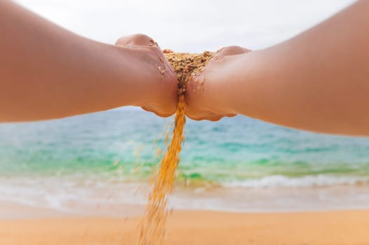 The hand releases the falling sand. Fine sand flows through your fingers against the backdrop of the sea. Summer beach holiday and time passing concept