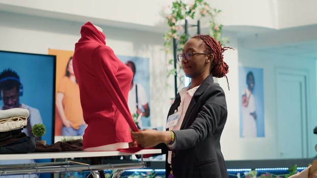 Meticulous retail assistant working in luxury fashion boutique, putting on women blouse on mannequin. Experienced african american employee showcasing new collection of trendy garments