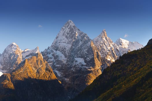 Sharp mountain peaks partially covered with snow and surrounded by rays of sun under a picturesque blue sky after dark, sunset or sunrise in the mountains in winter.