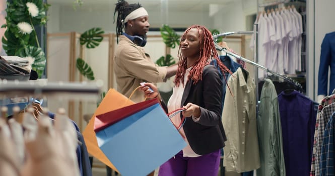 African american woman dancing in clothing store with shopping bags in hand, being happy about promotion sales. BIPOC customer in fashion boutique doing dance moves, being excited about low prices