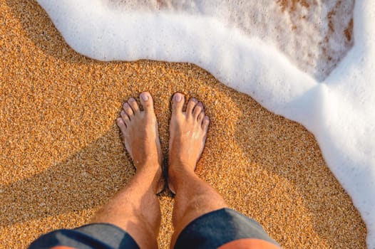 male feet barefoot on the waves of sea foam on a sandy golden beach on a summer day. top view of male legs in shorts and clear waters of the ocean.