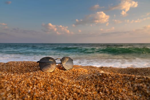 Sand dune and sunglasses. Sunglasses on clean sand with small bumps against the backdrop of the turquoise sea and sunset sky, close-up, side view.